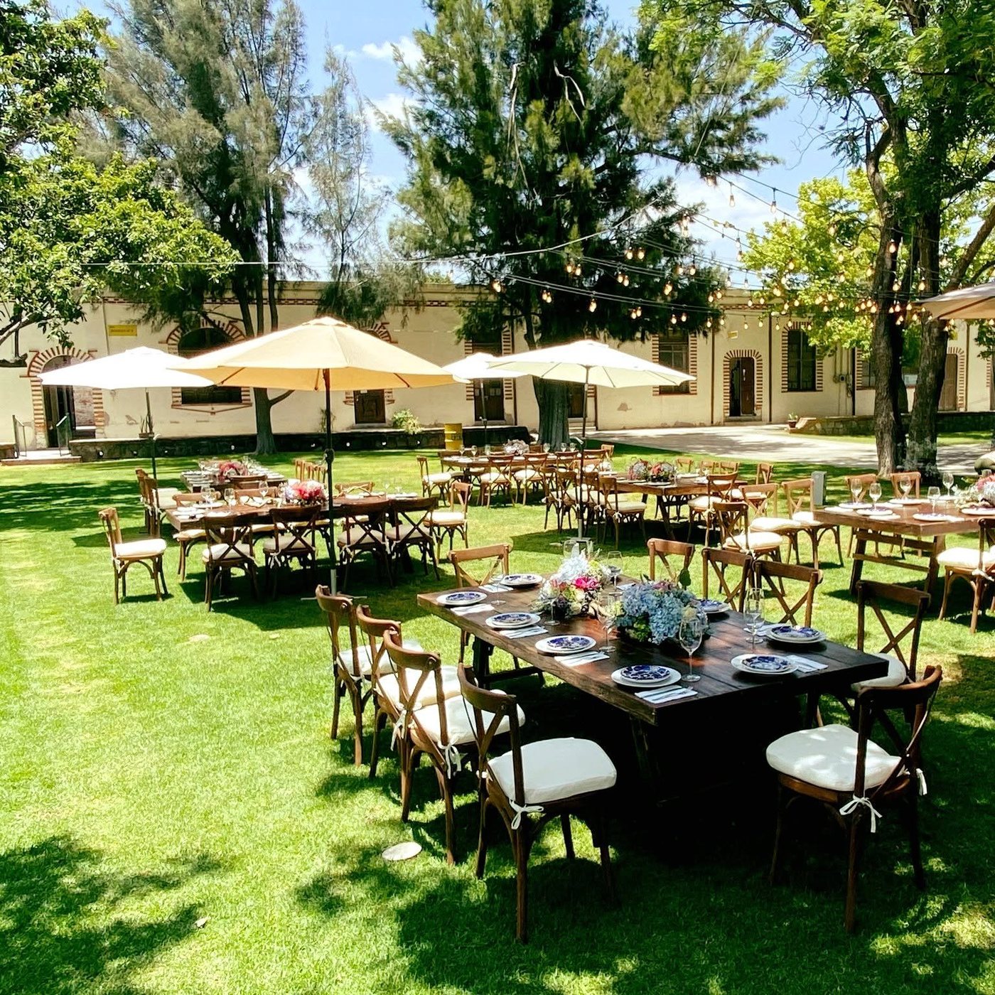 Jardín de día con mesas, sombrillas, árboles y la fachada de ladrillo al fondo